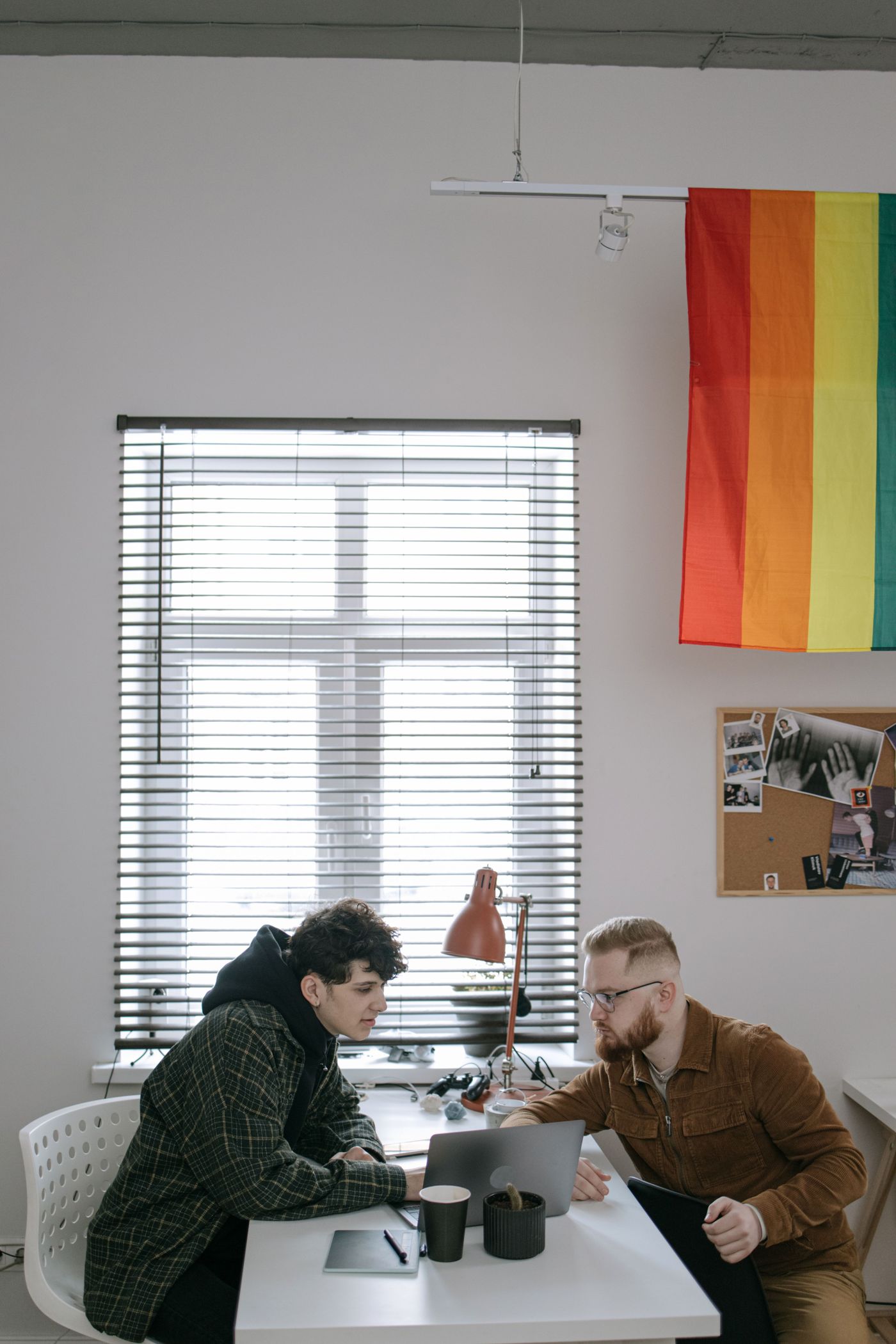 Deux personnes discutent devant un ordinateur dans un bureau, avec un drapeau arc-en-ciel au mur.